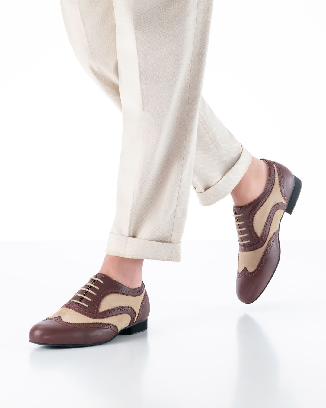 Un modelo masculino con pantalones de color crema camina hacia la cámara con zapatos de baile en tonos caramelo y burdeos, con diseño brogue perforado.