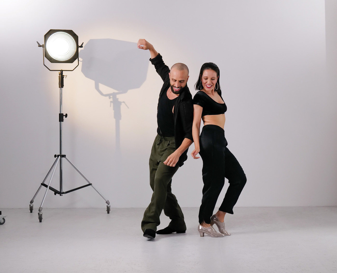 Movimientos dinámicos de bachata de una joven pareja de baile con atuendos oscuros y zapatos de baile de Werner Kern en plata y negro ante una pared blanca de estudio con un foco de luz al fondo.