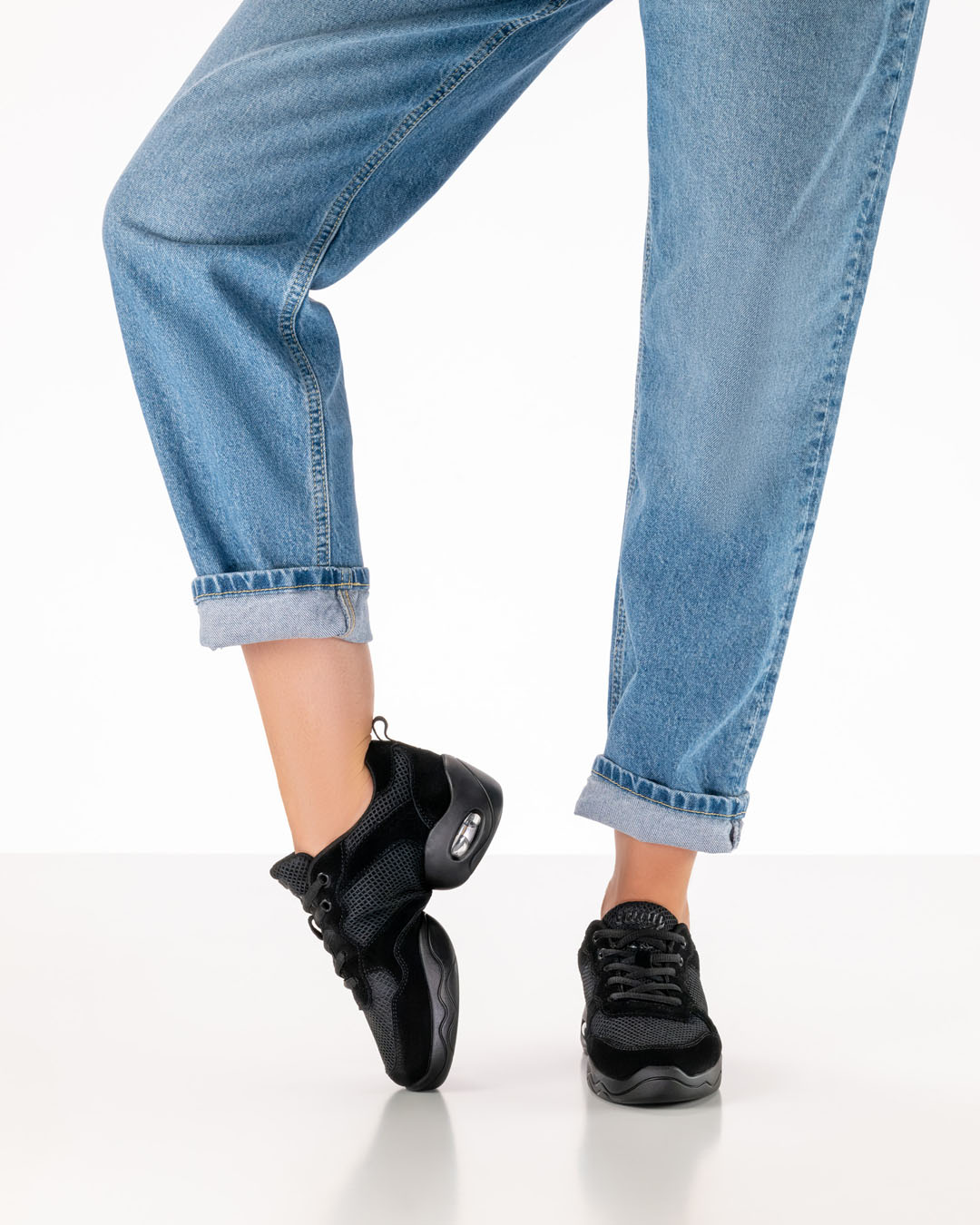 Una modelo vestida con vaqueros lleva unas zapatillas de baile negras con suela dividida y dobla el pie derecho por el empeine para mostrar la flexibilidad del calzado.